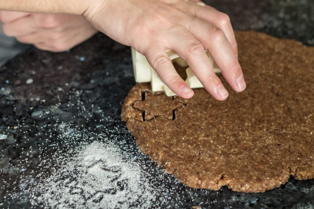 Eine Hand sticht Sternformen aus braunem Teig auf einer bemehlten Arbeitsfläche aus.