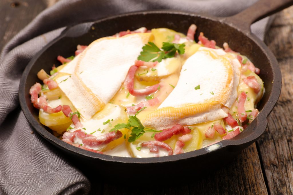 Tartiflette wird in einer schwarzen Pfanne mit Kartoffelscheiben, Speckstreifen und Käse überbacken.