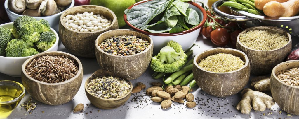 Verschiedene Schalen mit Vollkorngetreide, Hülsenfrüchten, Nüssen und Gemüse stehen dicht nebeneinander auf einem Tisch.