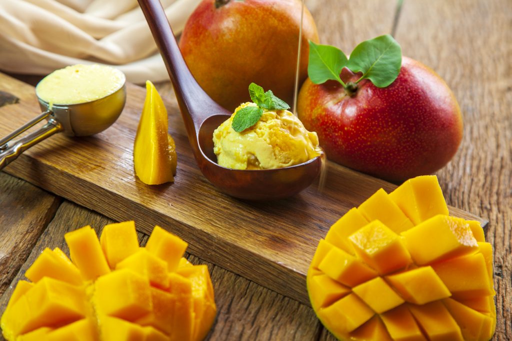 Ein Löffel Mangoeis liegt auf einem Holzbrett neben aufgeschnittener Mango und ganzen Früchten.