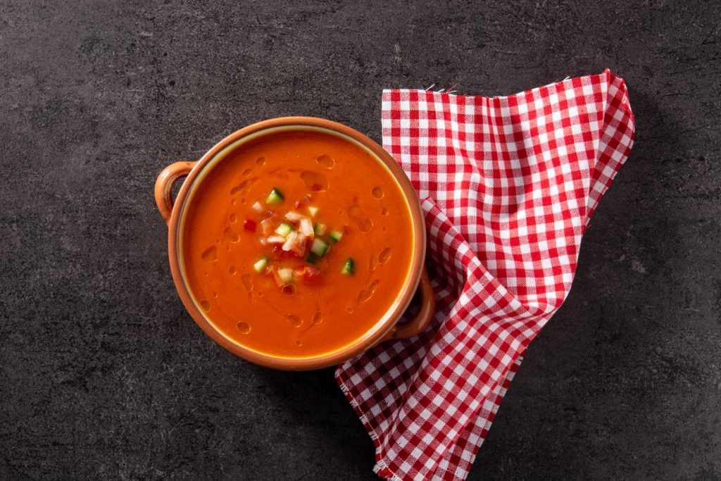 Aufnahme von oben einer Schale Gazpacho mit Gemüsewürfeln auf dunklem Untergrund neben einem rot weiß karierten Tuch.
