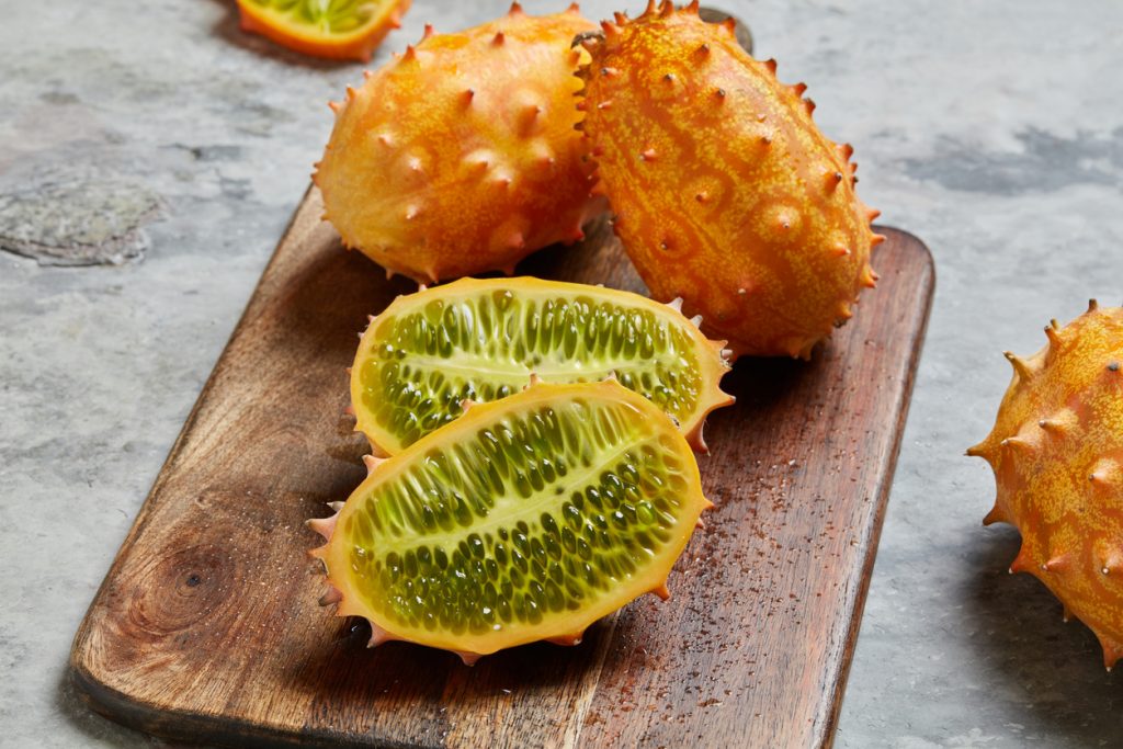 Zwei Kiwano-Hälften liegen auf einem Holzbrett, dahinter stehen weitere ganze Früchte auf grauem Untergrund.