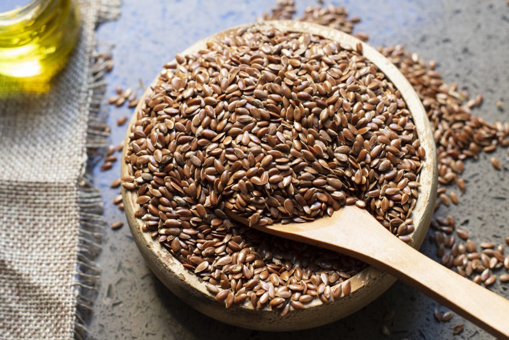 Eine Holzschale voller Leinsamen mit Holzlöffel liegt neben einem Glas Öl.