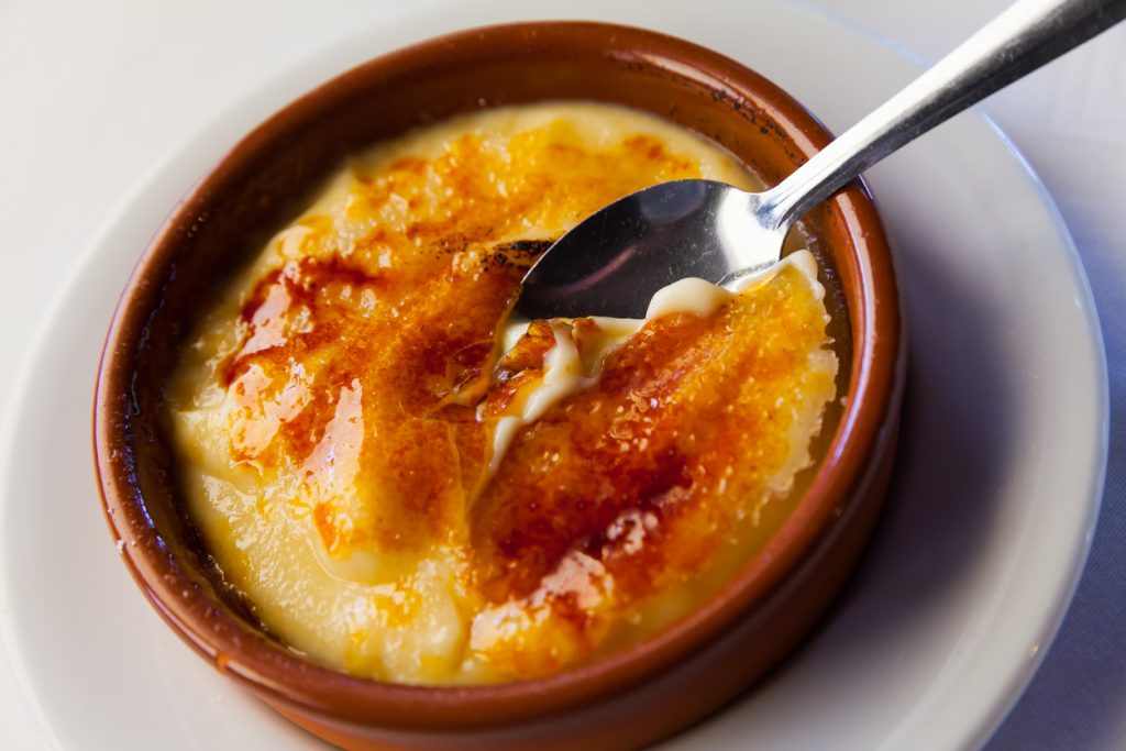 Ein Löffel sticht in die Crema Catalana und hebt ein Stück der knusprigen Zuckerkruste an.