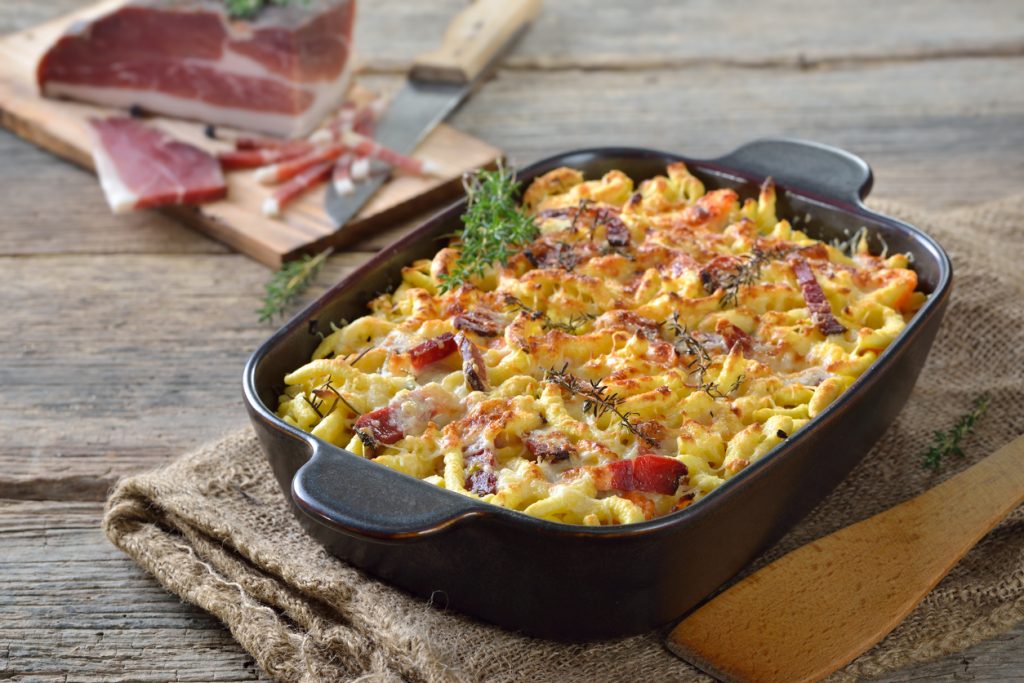 Eine Auflaufform mit Südtiroler Spätzleauflauf steht auf Jute neben Speck auf einem Brett.