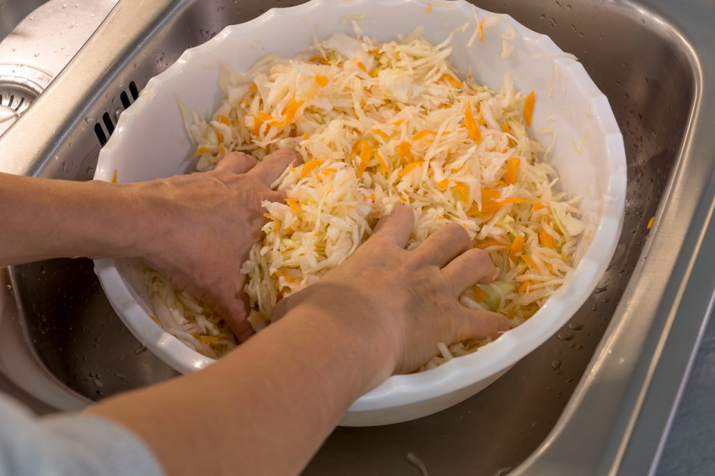 Hände kneten fein geschnittenen Weißkohl mit Karotten in einer Schüssel im Spülbecken.