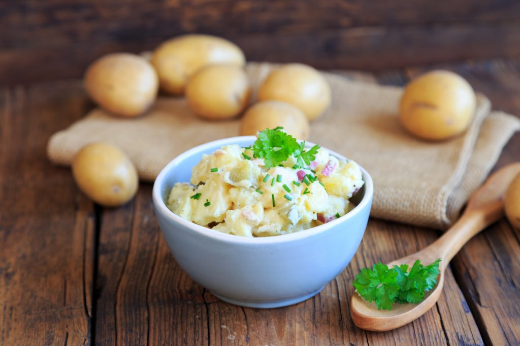 Eine Schüssel Kartoffelsalat mit Mayonnaise steht auf einem Holztisch, im Hintergrund liegen Kartoffeln.