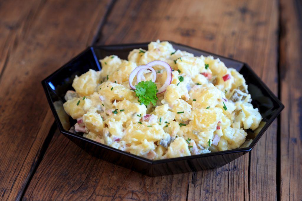 Ein Teller Kartoffelsalat mit Mayonnaise steht auf einem Holztisch und ist mit Kräutern und Zwiebelringen belegt.