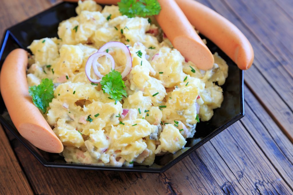 Kartoffelsalat mit Mayonnaise liegt auf einem schwarzen Teller und ist mit Kräutern und Zwiebelringen dekoriert.