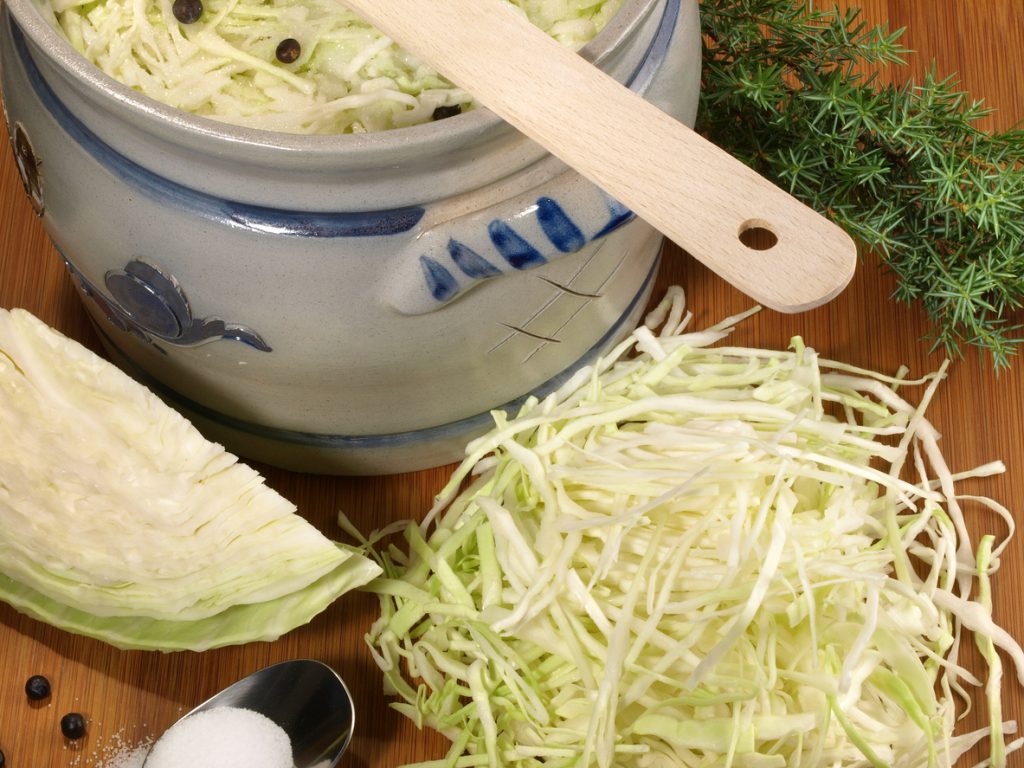 Ein Steinguttopf mit eingesalzenem Weißkohl steht neben Salz, Kohlstück und Gewürzen.