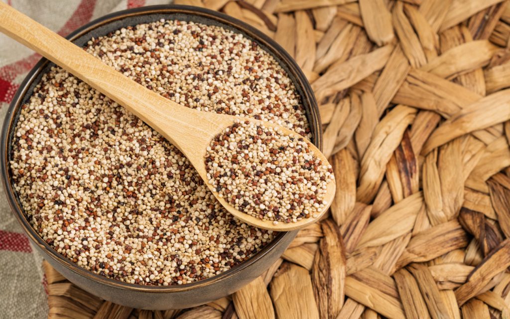 Eine Schale voller Quinoa mit einem Holzlöffel liegt auf einem geflochtenen Untergrund.