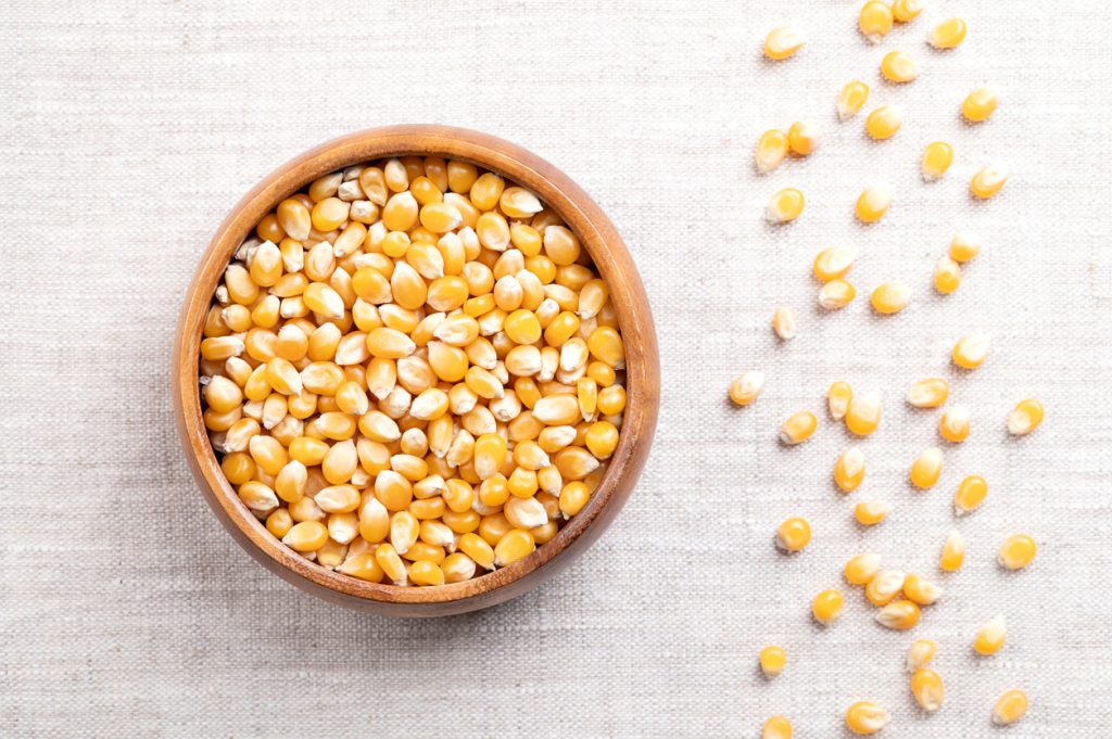 Gelbe Maiskörner liegen in einer Holzschale, daneben sind einzelne Körner auf hellem Stoff verstreut.