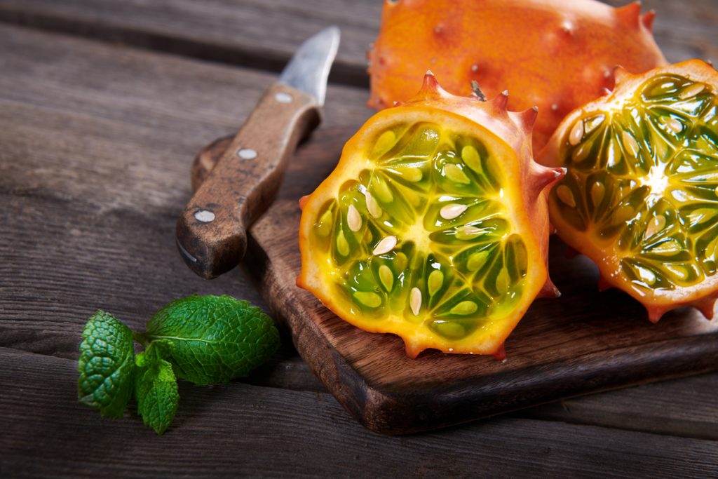 Eine halbierte Kiwano liegt auf einem Holzbrett neben einem Messer und Minze, das grüne Fruchtfleisch ist sichtbar.