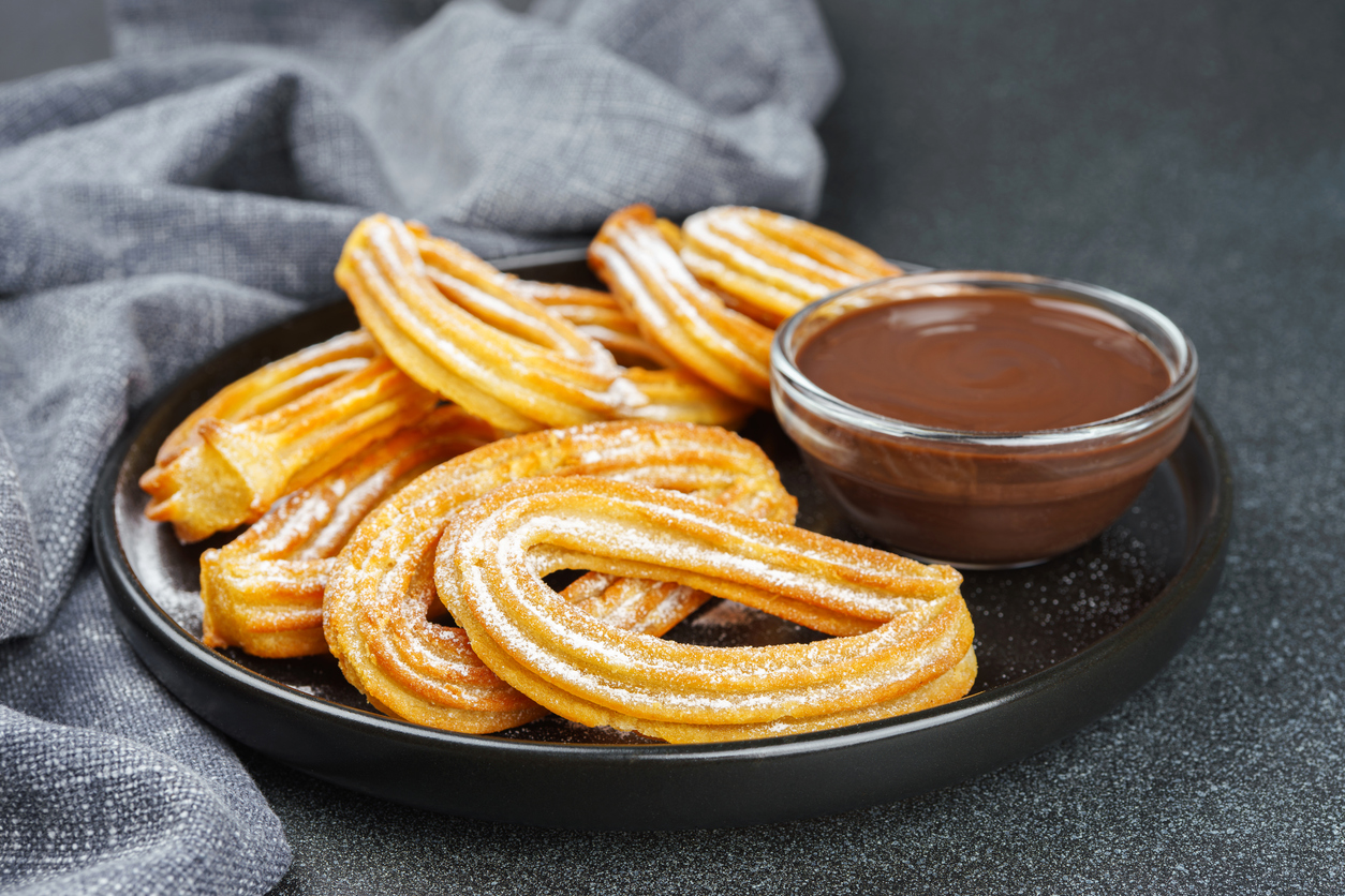 Zuckerbestäubte Churros auf schwarzem Teller mit Schokosauce in einer Glasschale.