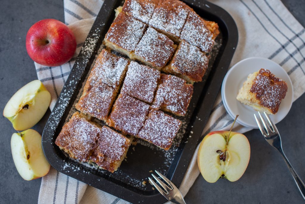 Ein Apfel-Zimt Kuchen liegt in Quadraten geschnitten auf einem Backblech neben Äpfeln.