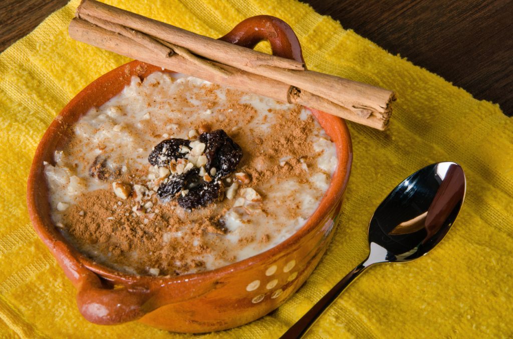Arroz con Leche steht in einem kleinen Tontopf auf einem gelben Tuch, oben liegen Rosinen, Nüsse und Zimtstangen.
