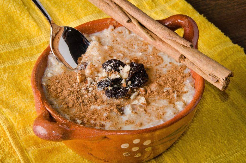 Ein Löffel steckt im Arroz con Leche, dazu liegen Zimtstangen auf dem Tontopf und Rosinen als Topping.