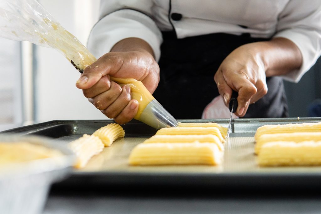 Koch spritzt Churros Teig mit Spritzbeutel in gleichmäßigen Streifen auf ein Blech.