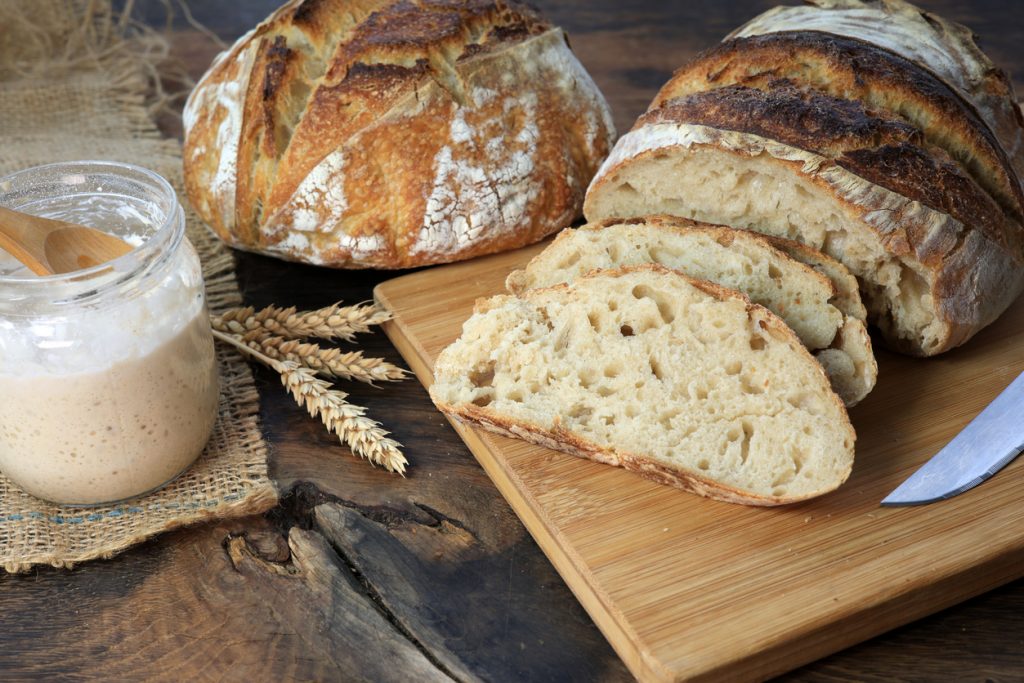 Ein Glas Sauerteigstarter steht neben geschnittenem Sauerteigbrot auf einem Holzbrett.