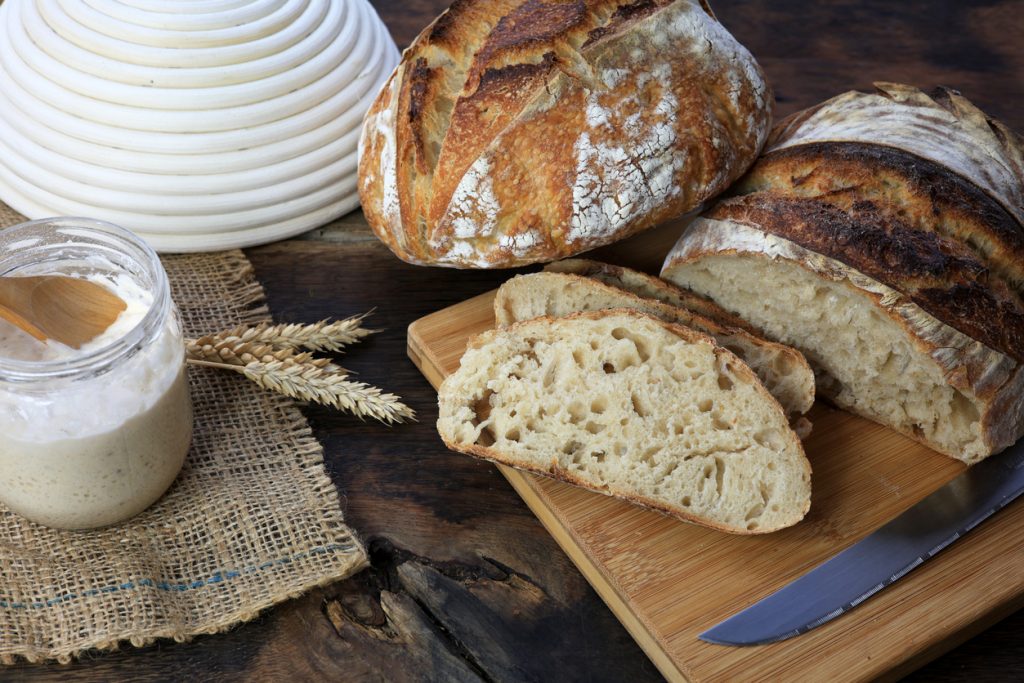 Sauerteigbrot und Brotscheiben liegen neben einem Glas Starter, einem Gärkorb und einem Messer auf dem Tisch.