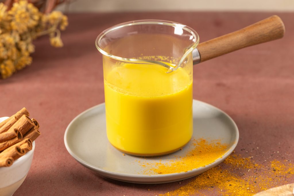 Ein Glasgefäß mit goldener Milch steht auf einem Teller neben Zimtstangen und Kurkumapulver.