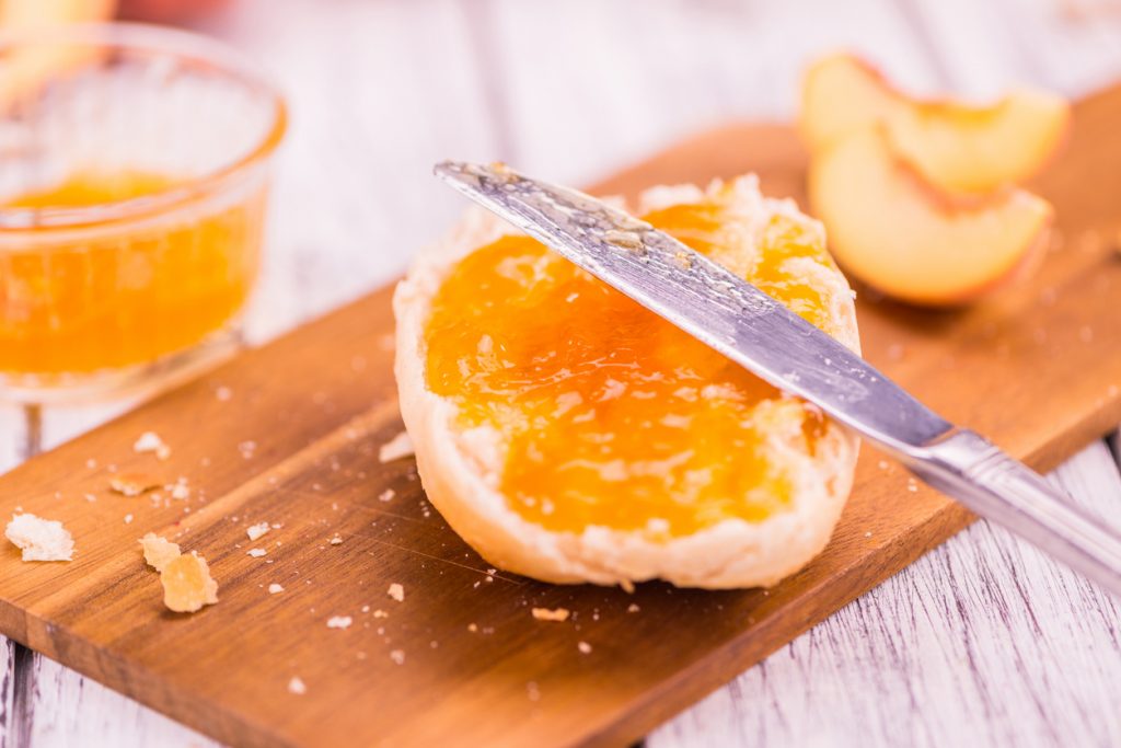 Ein aufgeschnittenes Brötchen ist mit Pfirsichmarmelade bestrichen und ein Messer liegt darüber.