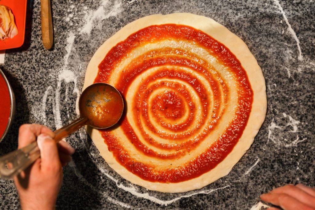 Eine Kelle verteilt Tomatensauce spiralförmig auf einem runden Pizzateig.