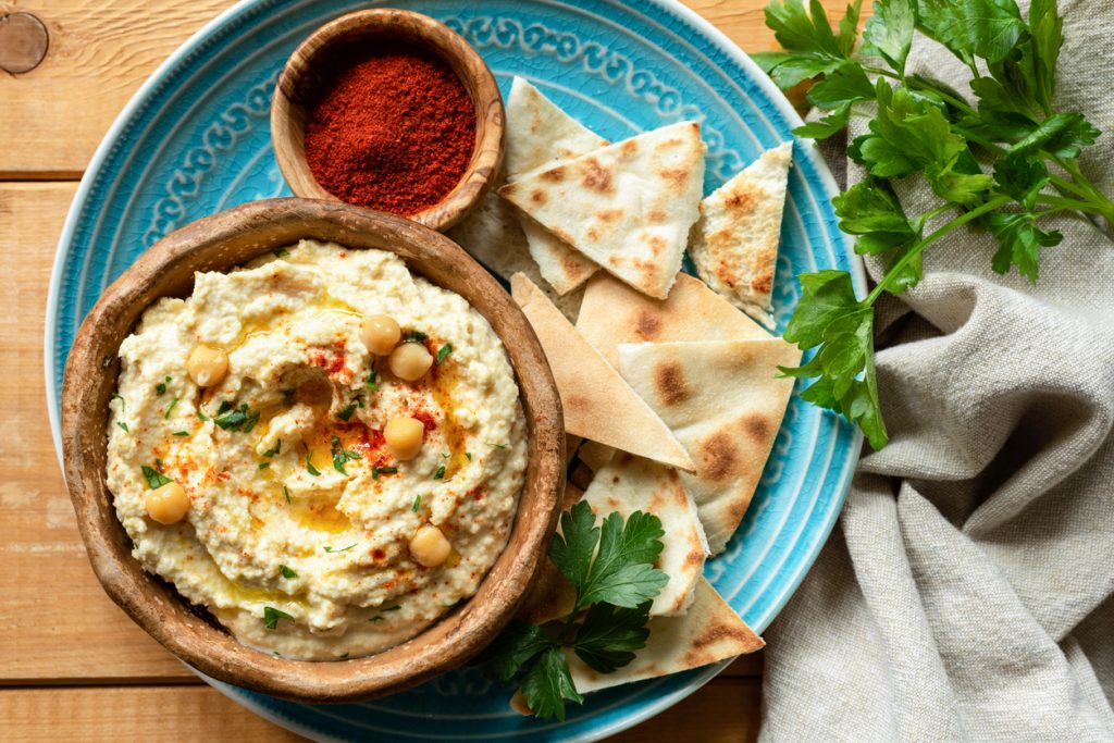 Eine Schale Hummus mit Kichererbsen und Öl steht auf einem blauen Teller neben Fladenbrot und Petersilie.