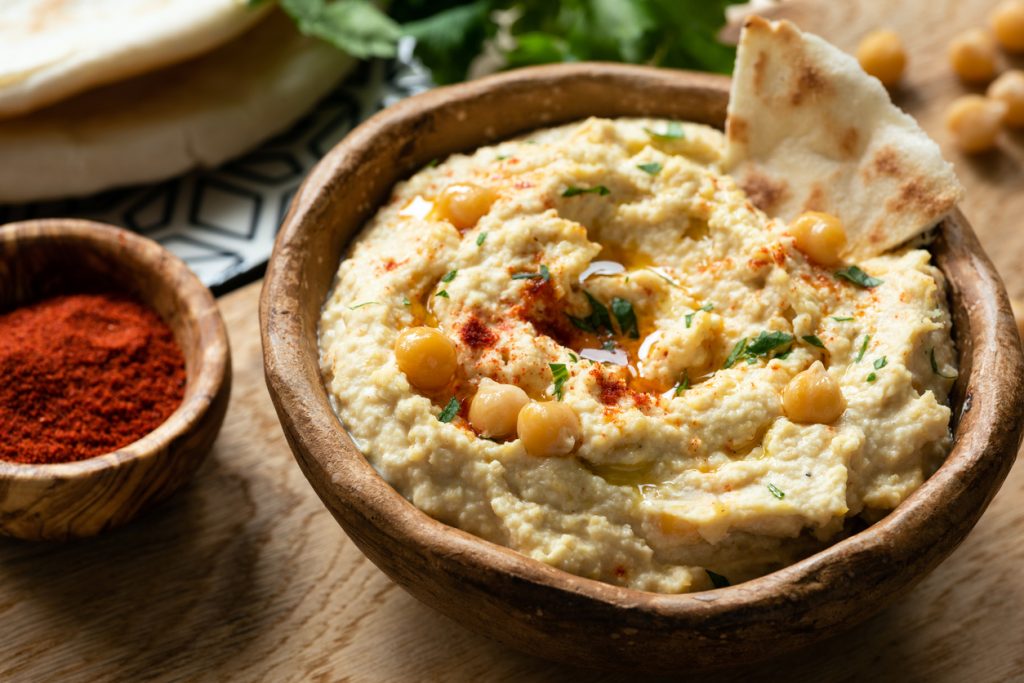 Hummus ist in einer Holzschale angerichtet und mit Kichererbsen, Gewürz und einem Stück Fladenbrot garniert.
