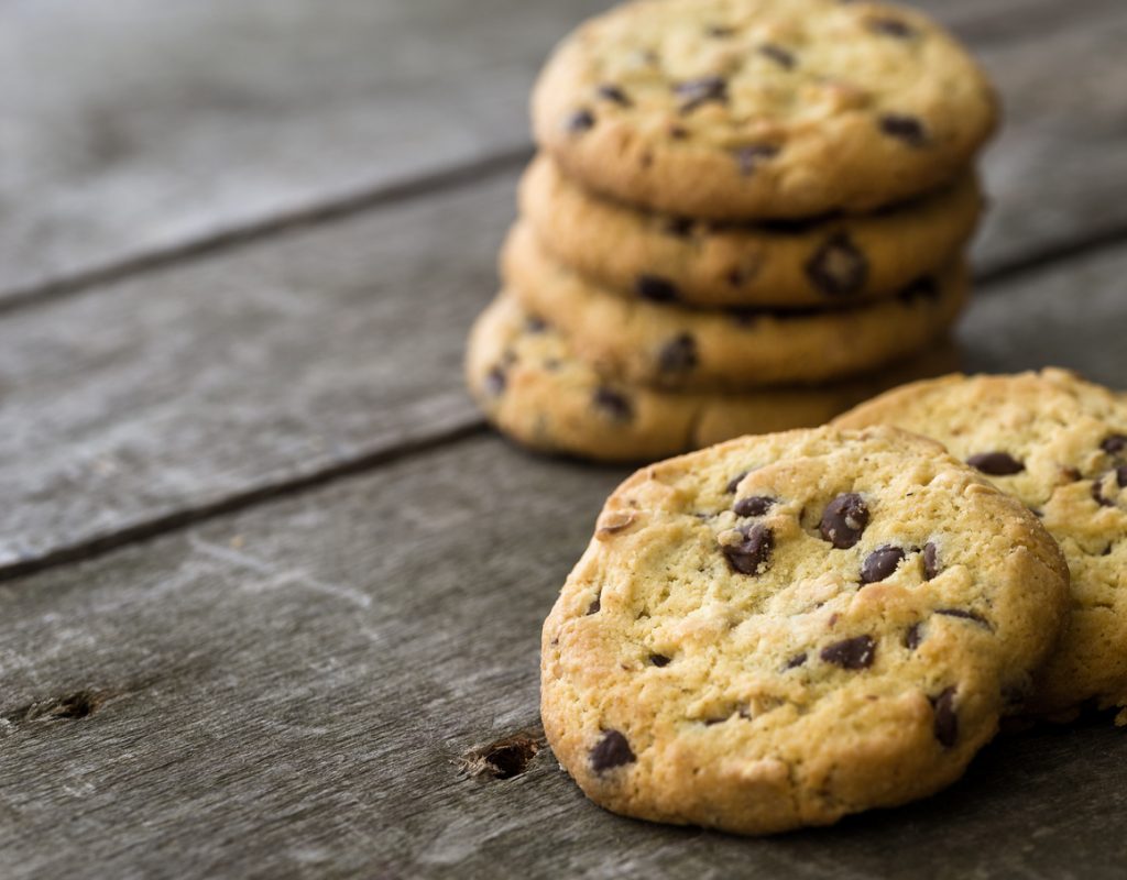 Ein Stapel Schoko-Cookies steht im Hintergrund, vorne liegt ein Cookie auf einem Holztisch.