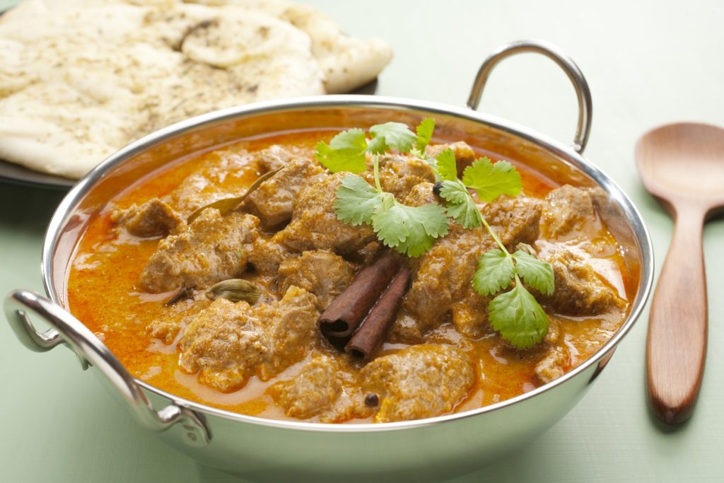 Rogan Josh Curry in einer großen Servierschale mit Naan im Hintergrund und Zimtstangen im Curry.