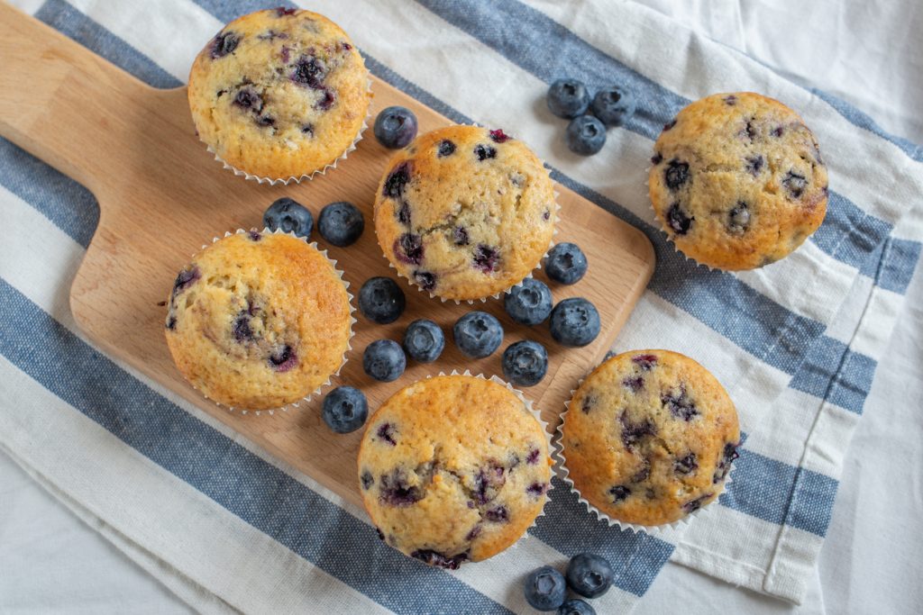 Mehrere Blaubeermuffins liegen auf einem Holzbrett, rundherum liegen Blaubeeren auf einem Tuch.