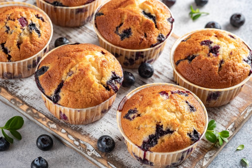 Mehrere Blaubeermuffins liegen auf einem Brett, daneben sind einzelne Blaubeeren verteilt.