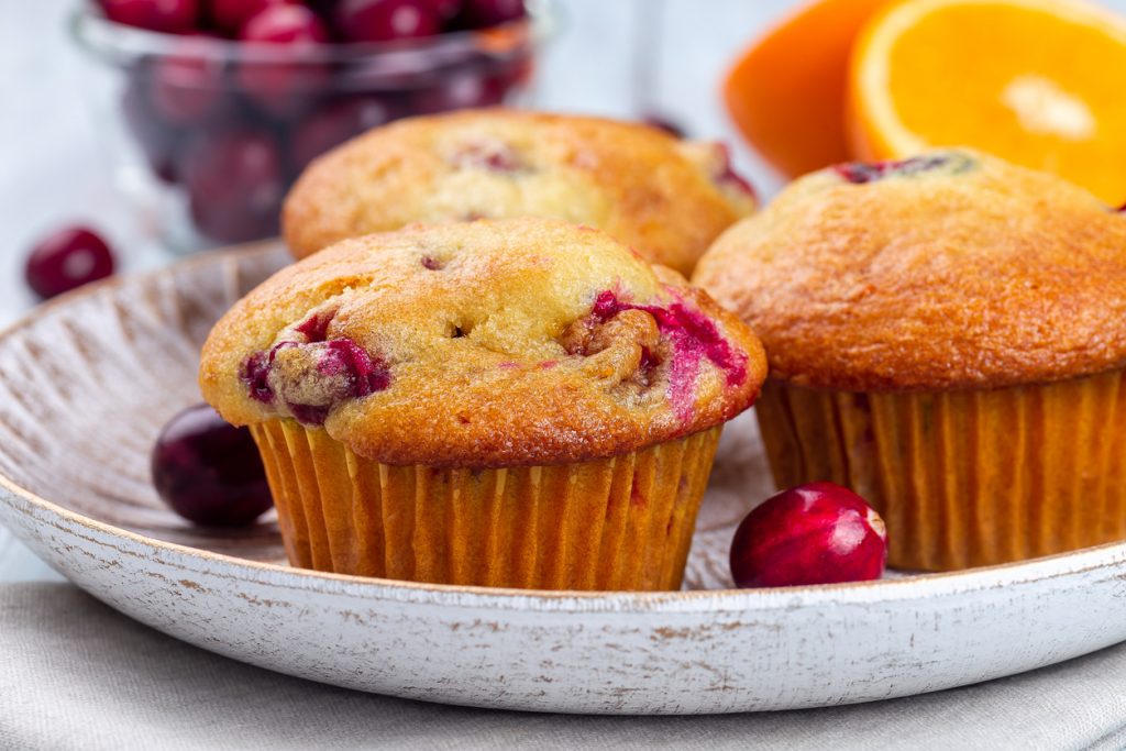 Drei Cranberry Muffins liegen auf einem Teller, daneben sind Cranberries und eine halbe Orange zu sehen.