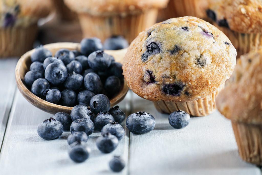 Ein Blaubeermuffin steht neben einer kleinen Schale mit Blaubeeren auf einem hellen Tisch.