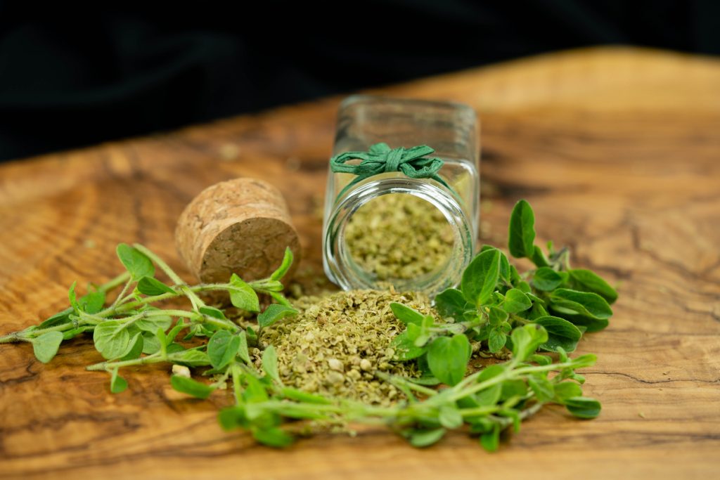 Getrockneter Oregano liegt vor einem offenen Glas, daneben liegen frische Oreganozweige auf Holz.
