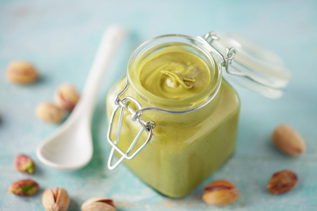 Ein kleines Glas mit grüner Pistazienbutter steht auf einem hellblauen Untergrund, umgeben von Pistazien.