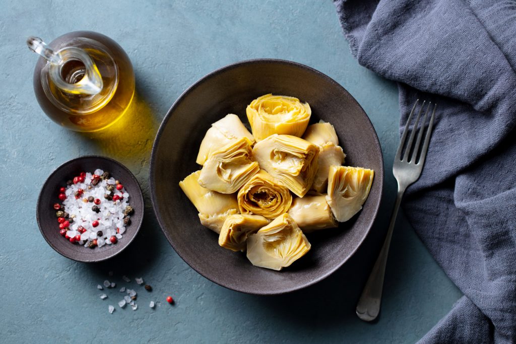 In einer Schale liegen Artischockenherzen, daneben stehen Olivenöl, grobes Salz und eine Gabel.
