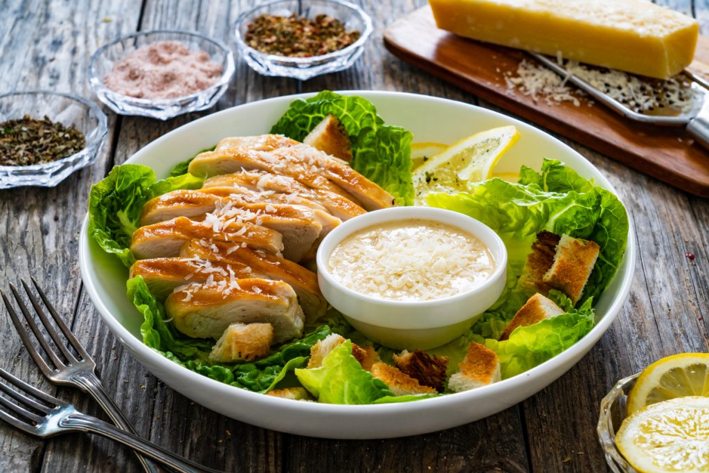 Ein Caesar Salat mit Hähnchenstreifen, Croutons und einer Schale Dressing ist auf einem Tisch angerichtet.