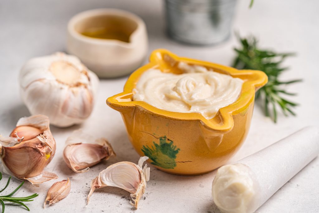 Ein Keramikschälchen mit cremiger Aioli steht auf einem hellen Tisch, daneben liegen Knoblauchzehen, Öl und Rosmarin.