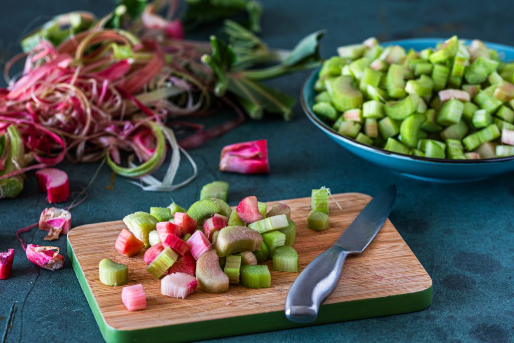 Geschnittener Rhabarber liegt auf einem Brett neben einem Messer, daneben steht eine Schüssel mit Stücken.