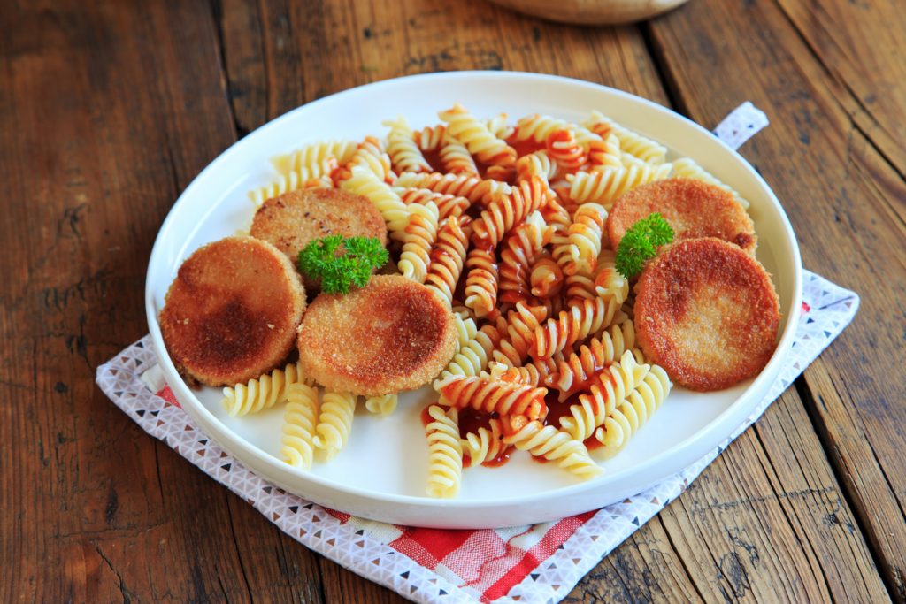 Ovaler Teller mit Spirelli in Tomatensoße und vier panierten Jagdwurstscheiben als DDR-Jägerschnitzel auf Holzuntergrund.