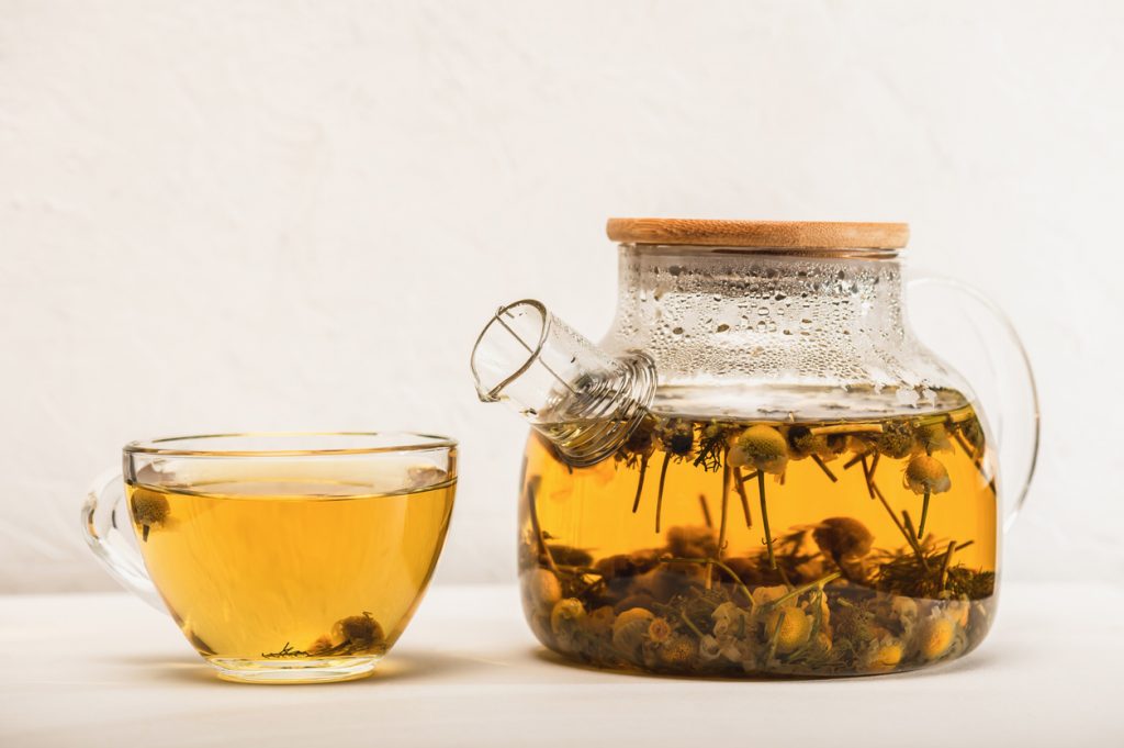 Eine Tasse Kamillentee steht neben einer Glaskanne mit Kamillenblüten im Tee.