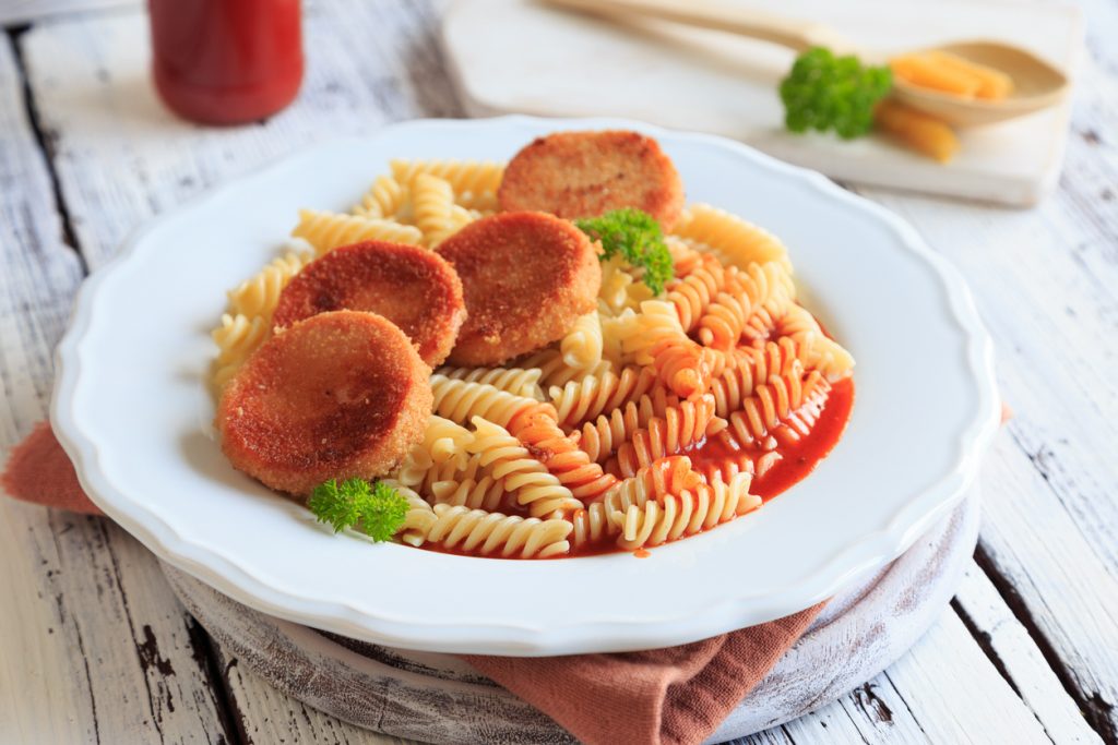 Vier panierte Jagdwurstscheiben liegen als DDR-Jägerschnitzel auf Spirelli mit Tomatensoße auf einem weißen Teller.