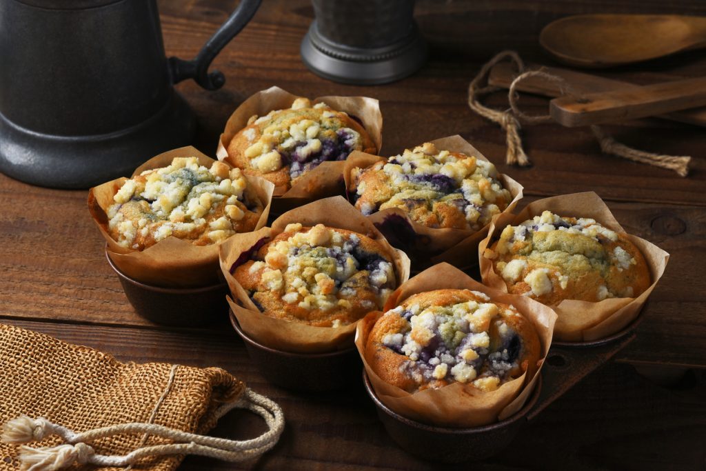 Mehrere Blaubeermuffins mit Streuseln stehen in Förmchen auf einem dunklen Holztisch.