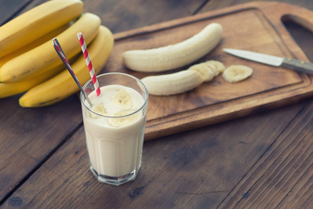 Ein Bananenshake im Glas mit zwei Trinkhalmen steht auf einem Holztisch neben Bananen und einem Schneidebrett.
