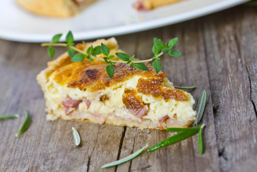 Ein Stück Quiche liegt auf einem Holztisch, Kräuterzweige sind darum verteilt.