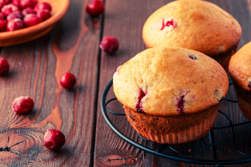 Ein Cranberry Muffin steht auf einem Gitter, während weitere Muffins und Cranberries auf einem Holztisch liegen.