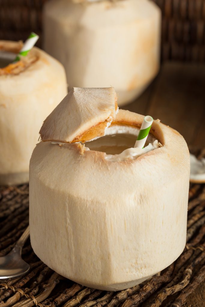 Eine geöffnete junge Kokosnuss steht mit Trinkhalm im warmen Licht, im Hintergrund sind weitere Kokosnüsse.