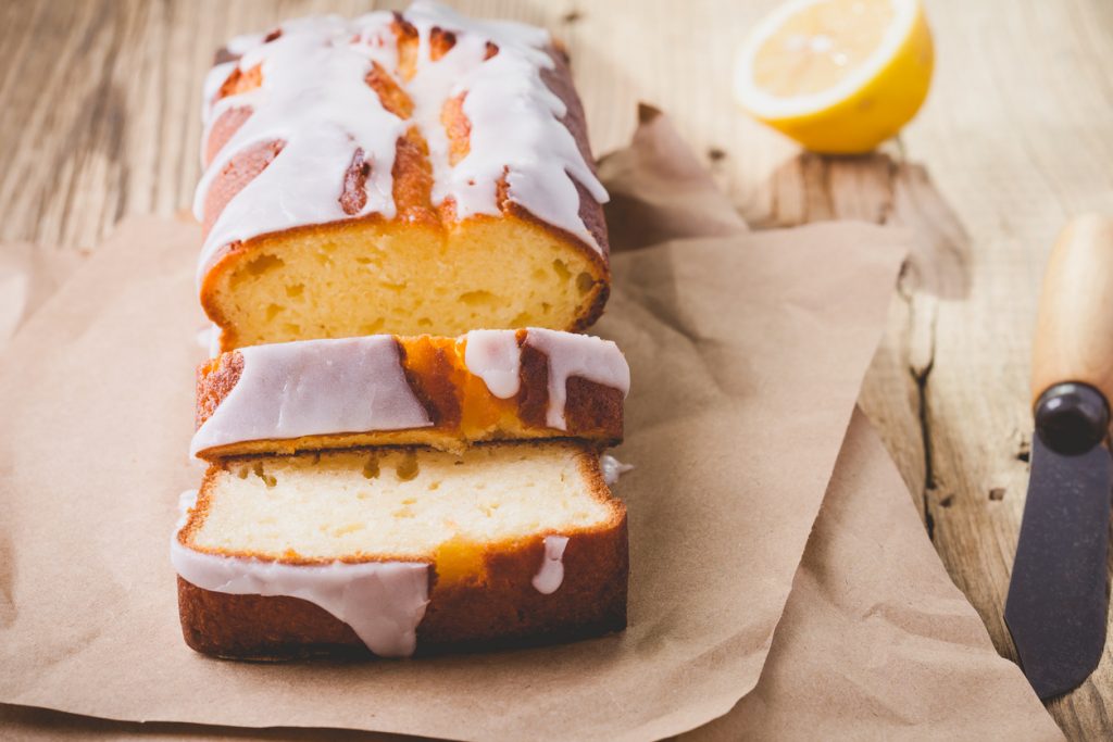 Ein angeschnittener Zitronenkuchen aus der Kastenform liegt auf Backpapier mit Zitrone im Hintergrund.